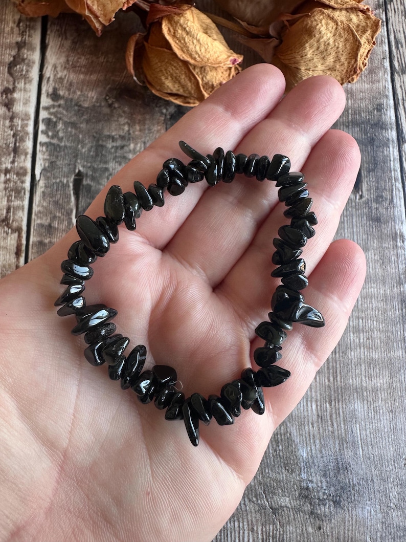 May include: A black gemstone bracelet composed of irregularly shaped, polished black stones. The bracelet is strung together, forming a circular shape. The bracelet is held in a person's hand, with a wooden background.