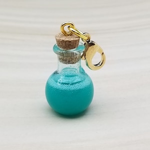 May include: A miniature glass potion bottle charm filled with a vibrant turquoise liquid. The bottle is sealed with a cork and has a gold-colored clasp. The charm is designed to be attached to a keychain or necklace.