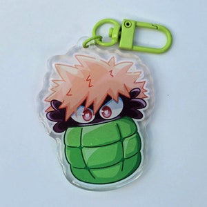 Puede incluir: Un llavero acrílico transparente con un personaje de dibujos animados con pelo naranja puntiagudo y ojos rojos. El personaje está sobre un fondo verde en forma de granada, con un cierre verde.
