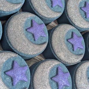 May include: Close-up of several round bath bombs. Each bath bomb has a blue base with a silver crescent moon and a purple star. The bath bombs are arranged in a grid pattern.