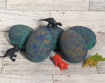 May include: Four green, blue, and gold speckled bath bombs shaped like dinosaur eggs.  The bath bombs are surrounded by small plastic dinosaur toys.