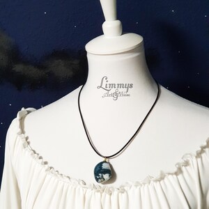 Puede incluir: Un collar de cord&oacute;n negro con un colgante de piedra azul y blanca. El colgante, de forma ovalada, tiene detalles dorados. El collar se exhibe en un maniqu&iacute; blanco con una blusa blanca.