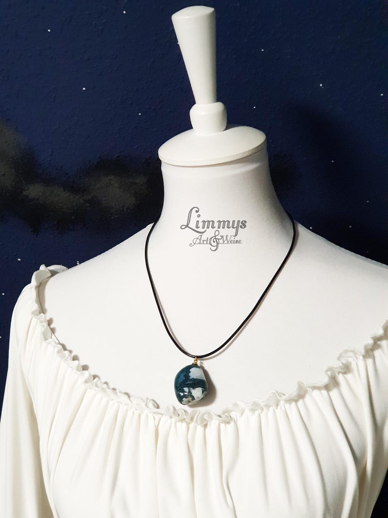 Puede incluir: Un collar de cord&oacute;n negro con un colgante de piedra azul y blanca. El colgante, de forma ovalada, tiene detalles dorados. El collar se exhibe en un maniqu&iacute; blanco con una blusa blanca.
