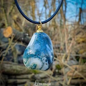 Puede incluir: Un colgante de piedra azul y blanca con una tapa dorada cuelga de un cord&oacute;n negro. La piedra tiene un aspecto veteado. El colgante se exhibe al aire libre con un fondo borroso de &aacute;rboles y follaje.