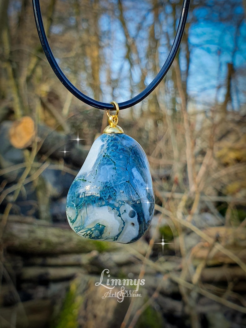 Puede incluir: Un colgante de piedra azul y blanca con una tapa dorada cuelga de un cord&oacute;n negro. La piedra tiene un aspecto veteado. El colgante se exhibe al aire libre con un fondo borroso de &aacute;rboles y follaje.