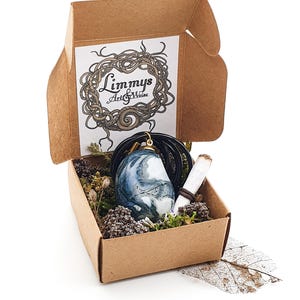 Puede incluir: Una peque&ntilde;a caja de cart&oacute;n abierta contiene un colgante de piedra veteada azul y blanca sobre un cord&oacute;n negro, un papel enrollado y plantas secas. La caja tiene la inscripci&oacute;n "Limmys Art & Weise".
