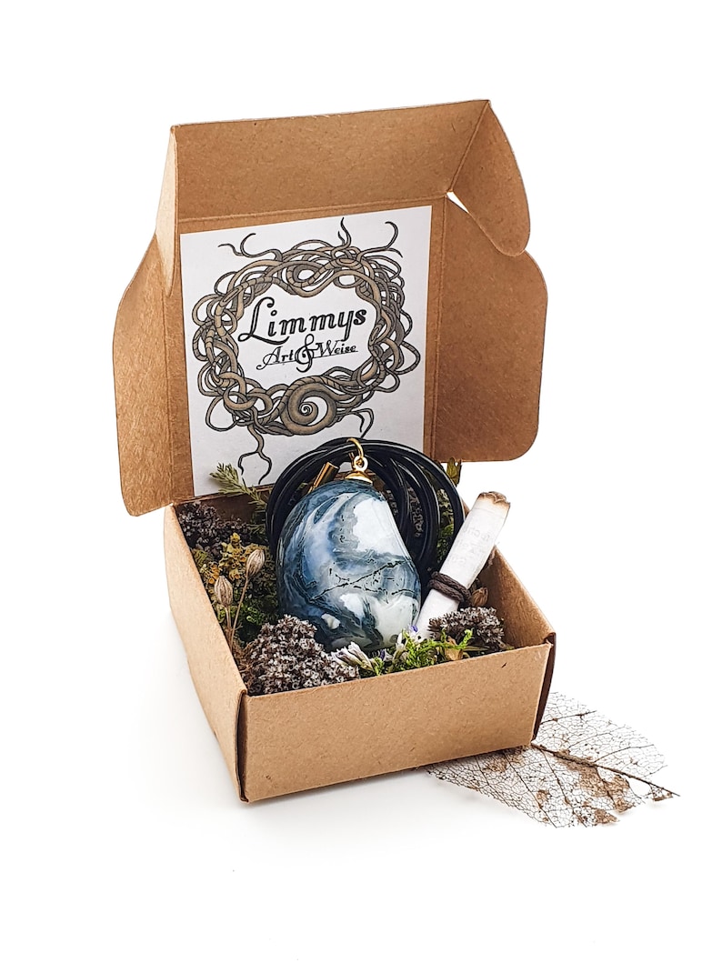 Puede incluir: Una peque&ntilde;a caja de cart&oacute;n abierta contiene un colgante de piedra veteada azul y blanca sobre un cord&oacute;n negro, un papel enrollado y plantas secas. La caja tiene la inscripci&oacute;n "Limmys Art & Weise".