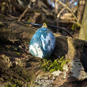 Puede incluir: Colgante ovalado de piedra azul y blanca con una fianza dorada, suspendido de un cord&oacute;n negro. El colgante descansa sobre una superficie de madera texturizada y cubierta de musgo. El logo "Limmys Art & More" es visible.