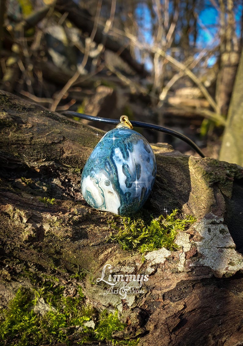 Puede incluir: Colgante ovalado de piedra azul y blanca con una fianza dorada, suspendido de un cord&oacute;n negro. El colgante descansa sobre una superficie de madera texturizada y cubierta de musgo. El logo "Limmys Art & More" es visible.