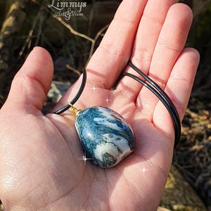 Puede incluir: Un collar con colgante que presenta una piedra grande y pulida con un patr&oacute;n veteado de color azul, verde y blanco. La piedra est&aacute; suspendida de un cord&oacute;n negro con una fianza dorada. El collar se sostiene en una mano.