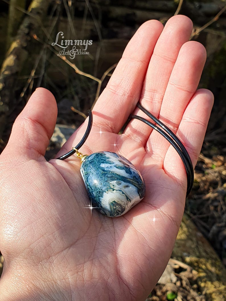 Puede incluir: Un collar con colgante que presenta una piedra grande y pulida con un patr&oacute;n veteado de color azul, verde y blanco. La piedra est&aacute; suspendida de un cord&oacute;n negro con una fianza dorada. El collar se sostiene en una mano.