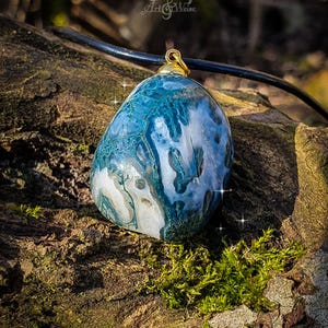 Puede incluir: Colgante de piedra azul y blanca veteada, con una fianza dorada, suspendido de un cord&oacute;n negro. La piedra presenta un patr&oacute;n &uacute;nico, sobre un fondo natural y musgoso. Probablemente una pieza de joyer&iacute;a hecha a mano.