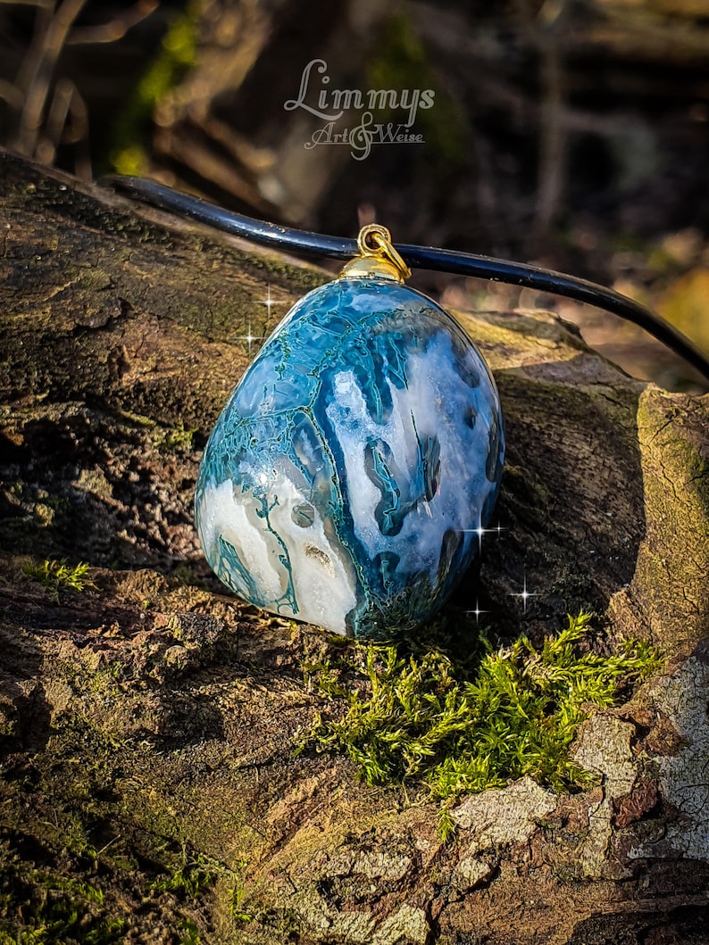 Puede incluir: Colgante de piedra azul y blanca veteada, con una fianza dorada, suspendido de un cord&oacute;n negro. La piedra presenta un patr&oacute;n &uacute;nico, sobre un fondo natural y musgoso. Probablemente una pieza de joyer&iacute;a hecha a mano.