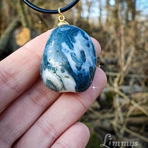 Puede incluir: Un colgante de piedra de &aacute;gata azul y blanca con una fianza dorada, suspendido de un collar de cord&oacute;n negro. La piedra tiene un aspecto veteado con patrones arremolinados. El colgante se sostiene en una mano sobre un fondo natural borroso.