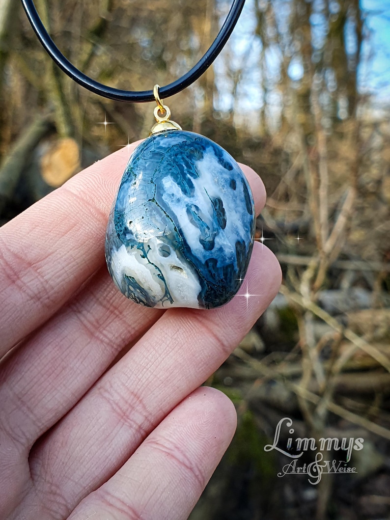 Puede incluir: Un colgante de piedra de &aacute;gata azul y blanca con una fianza dorada, suspendido de un collar de cord&oacute;n negro. La piedra tiene un aspecto veteado con patrones arremolinados. El colgante se sostiene en una mano sobre un fondo natural borroso.