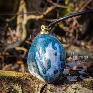 Puede incluir: Un colgante de &aacute;gata azul y blanca con una fianza dorada, suspendido de un cord&oacute;n negro. La piedra tiene un aspecto veteado con tonos de azul, blanco y toques dorados. El colgante se muestra sobre un trozo de madera.