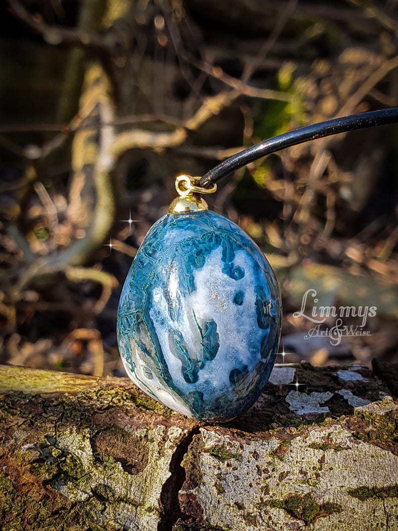 Puede incluir: Un colgante de &aacute;gata azul y blanca con una fianza dorada, suspendido de un cord&oacute;n negro. La piedra tiene un aspecto veteado con tonos de azul, blanco y toques dorados. El colgante se muestra sobre un trozo de madera.