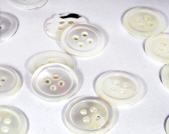 May include: A collection of round, white, four-hole buttons with a smooth, iridescent finish. Each button features a slightly recessed center and a raised outer rim. The buttons are scattered across the frame, with one button highlighted in a small inset box.