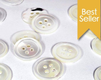 May include: A collection of white, round, four-hole buttons made of mother of pearl. The buttons are arranged on a white surface.