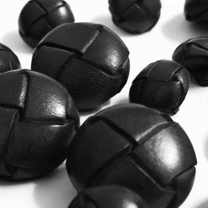 May include: A close-up of black leather buttons with a woven design. The buttons are arranged in a scattered pattern on a white surface.