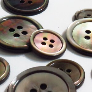 May include: A collection of 10 iridescent brown mother of pearl buttons with four holes each. The buttons are various sizes and are arranged in a random pattern on a white surface.