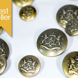 May include: A group of 10 gold-toned metal buttons with a raised design of a crest featuring a crown, two lions, and a banner. The buttons are scattered on a white surface.