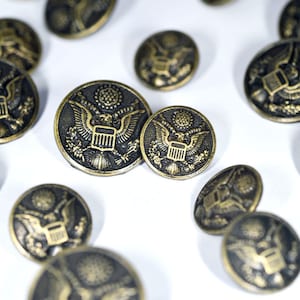 Peut inclure: Gros plan d'un groupe de boutons dorés anciens avec un motif d'aigle et de bouclier. Les boutons sont éparpillés sur une surface blanche.