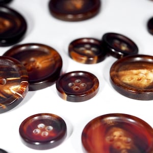 Puede incluir: Una colección de botones marrones y negros con un patrón moteado. Los botones tienen cuatro agujeros cada uno y están hechos de un material natural.