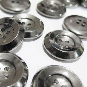 May include: A close-up of a group of silver metal buttons with four holes each. The buttons are arranged in a random pattern on a white surface.