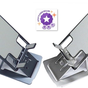 Puede incluir: Dos soportes para teléfono de metal plateado con una superficie texturizada. Los soportes están en posición plegada, listos para ser utilizados para sostener un teléfono.