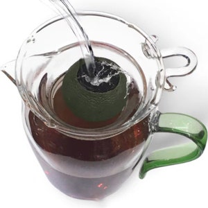 Pode incluir: Uma jarra de vidro com uma pega verde e uma tampa transparente. A jarra está cheia de um líquido escuro e um abacate verde está submerso no líquido.