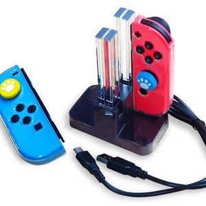 Op de afbeelding: Een zwart oplaadstation met twee transparante slots voor het opladen van Nintendo Switch Joy-Cons. Een rode Joy-Con met een blauwe pootprintknop zit in een slot. Een blauwe Joy-Con met een gele pootprintknop ligt op de tafel naast het oplaadstation.