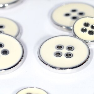May include: A set of five white buttons with silver trim and four holes each. The buttons are arranged in a semi-circle on a white surface.