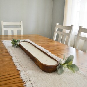 A large brown dough bowl candle unlit sitting on a white table runner with greenery