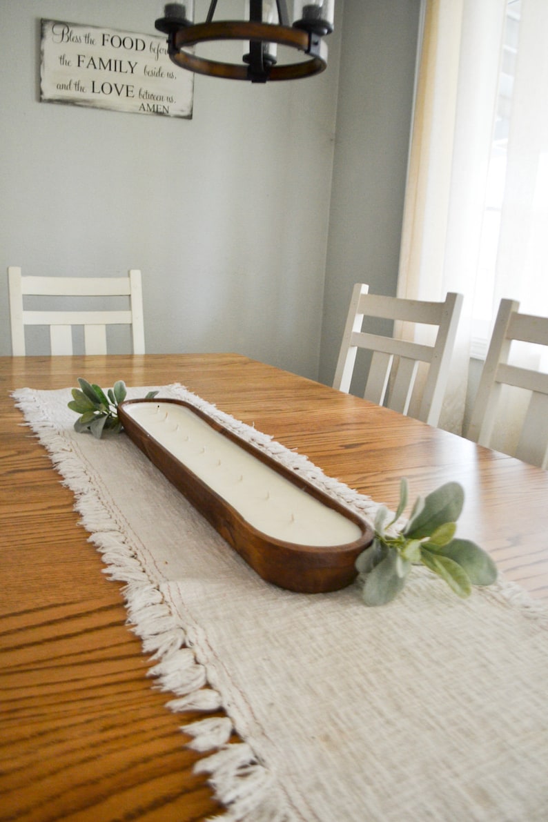 A large brown dough bowl candle unlit sitting on a white table runner with greenery