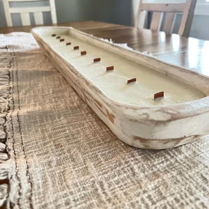 A white dough bowl candle unlit with wooden wicks sitting on a white table runner
