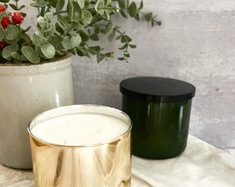 Puede incluir: Dos velas perfumadas en recipientes de vidrio. Una vela tiene un acabado metálico dorado, la otra es verde oscuro con una tapa negra. Una maceta de cerámica con vegetación y bayas rojas está en el fondo.