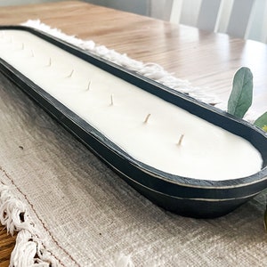 A black dough bowl candle with cotton wicks unlit sitting on a white table runner with faux flowers tucked under each end of the candle