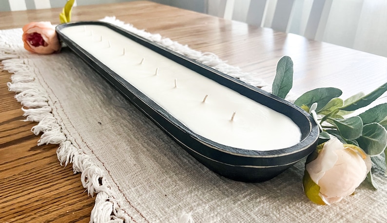 A black dough bowl candle with cotton wicks unlit sitting on a white table runner with faux flowers tucked under each end of the candle