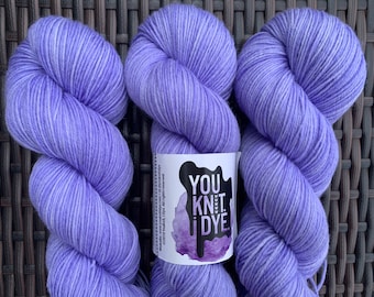 Può includere: Sono mostrati tre gomitoli di filato color lavanda, ciascuno ben avvolto e strutturato. È visibile un'etichetta bianca con la scritta "YOU KNIT i DYE". Il filato è disposto su una superficie tessuta marrone scuro.