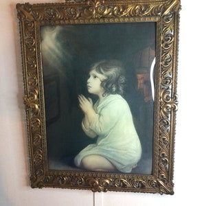 Puede incluir: Una impresión enmarcada de una niña arrodillada en oración con una luz brillando sobre ella. El marco es ornamentado y de color dorado.