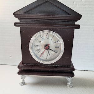 May include: A dark brown wooden mantel clock with a silver-colored clock face. The clock has Roman numerals and a red second hand. The clock sits on four silver-colored claw feet.