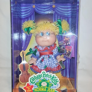 May include: A Cabbage Patch Kids Norma Jean doll in a box. The doll has blonde pigtails with green bows, glasses, and a blue and white checkered dress. The box features a stage backdrop and the text "Star of the Videos!" and "Special Edition".