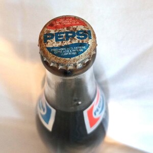 May include: A vintage glass bottle of Pepsi Cola with a rusty metal cap. The cap has the Pepsi logo and the ingredients listed: phosphoric acid, caffeine, citric acid, and natural flavorings.