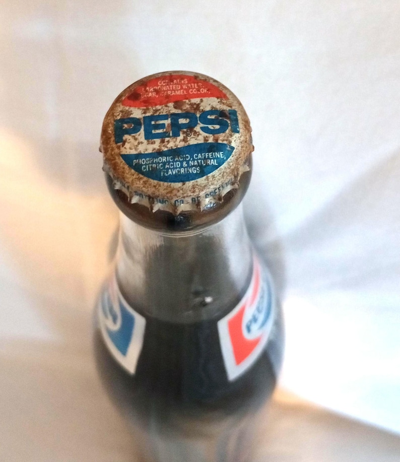 May include: A vintage glass bottle of Pepsi Cola with a rusty metal cap. The cap has the Pepsi logo and the ingredients listed: phosphoric acid, caffeine, citric acid, and natural flavorings.