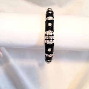 May include: A black enamel bangle bracelet with silver-tone accents. The bracelet features a repeating pattern of silver-tone filigree and clear, round gemstones. The bracelet is a classic piece of jewelry.