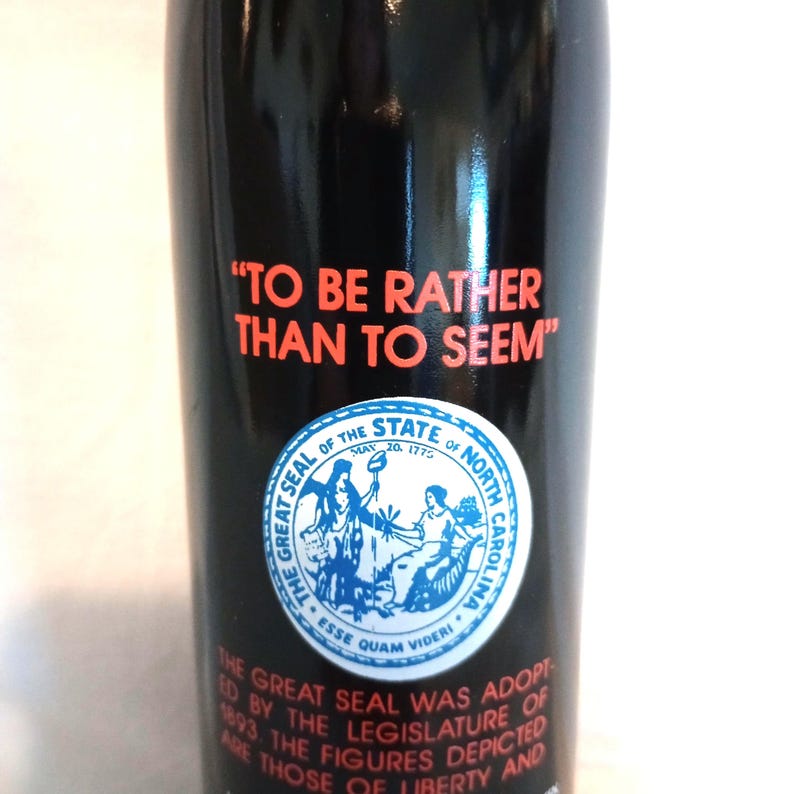 May include: A black bottle with a red and white label that says "TO BE RATHER THAN TO SEEM". The label also features the Great Seal of the State of North Carolina, which is a blue circle with a white star and the words "Esse Quam Videri".