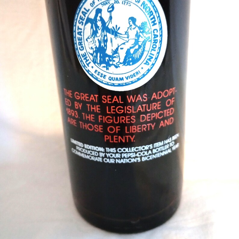 May include: A black glass bottle with a red label featuring the Great Seal of North Carolina. The label includes the text "The Great Seal was adopted by the Legislature of 1893. The figures depicted are those of Liberty and Plenty."