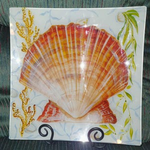 May include: Square glass decorative plate featuring a large, detailed seashell in shades of orange and white. The plate has a light blue background with painted seaweed and coral accents. The plate is supported by a black metal stand.