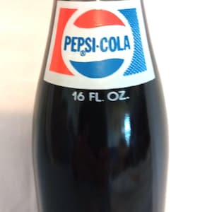 May include: A glass bottle of Pepsi-Cola with a red, white, and blue label. The bottle is 16 fluid ounces and has the quote "To be rather than to seem" printed on the bottom.